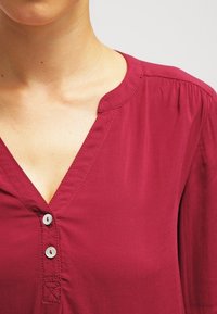 Blusa burdeos con escote en V, que presenta un cierre de botones y sutiles pliegues en los hombros, confeccionada con una tela suave y ligera.