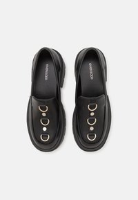 Paire de mocassins en cuir noir avec semelles épaisses, dotée de trois anneaux métalliques et de deux clous sur chaque bride supérieure, portant la marque "Even&Odd" à l'intérieur.