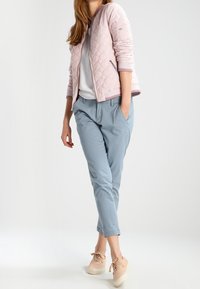 Frau trägt eine hellrosa gesteppte Jacke, ein weißes Oberteil, blaue Cropped-Hosen und beige Sneakers und steht mit einer Hand in der Tasche.