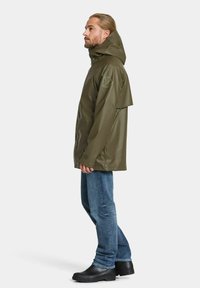 Olivgrön vattentät jacka med huva, med en något lös passform, synliga sömmar och en emblem på vänster ärm. Kombinerad med blå jeans och svarta stövlar.