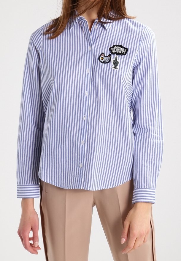 Femme portant une chemise à rayures verticales bleu et blanc avec des patchs de chat et de cactus et un pantalon beige.