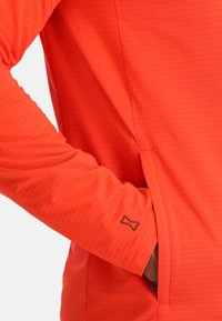 Langdurige mouw, fel oranje top. Heeft een gestructureerd patroon, zijzak en een klein accentlogo op de mouw. Glad materiaal.