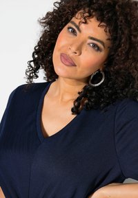 Ciemnoniebieska bluzka w serek z prążkowanej tkaniny, krótki rękaw i luźny krój. Duże srebrne kolczyki w kształcie kół podkreślają całość.