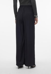 Zwarte wijde broek van textuurstof, met een elastische tailleband en subtiele plooien die naar de grond stromen. Gemaakt in combinatie met een getailleerd topje en hakken.