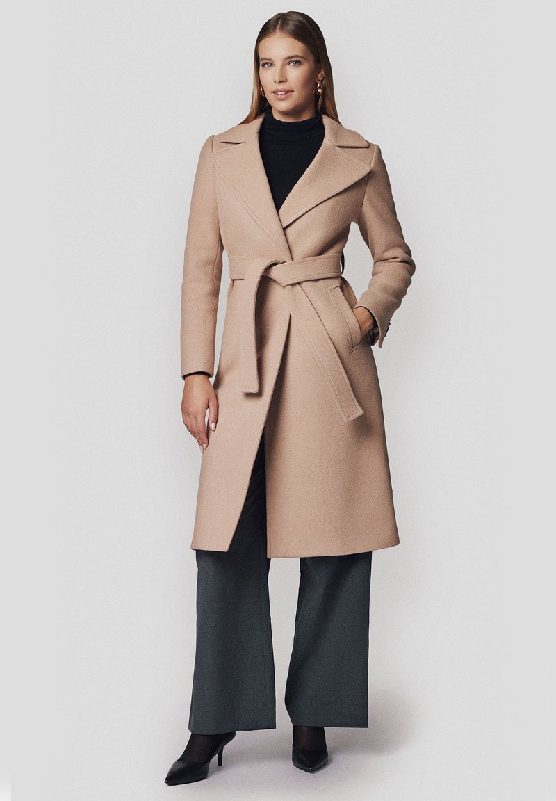 Beżowy płaszcz z wełny z szerokim kołnierzem, z wiązaniem, z paskiem do samodzielnego zawiązania i długimi rękawami; noszony na czarnym golfie i szarych spodniach.