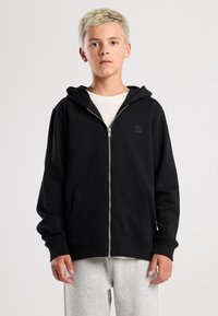 Schwarze Zip-up-Hoodie aus Baumwollmischung, in einfarbiger Ausführung, mit vorderen Taschen und gefütterter Kapuze. Über einem hellen Shirt getragen.