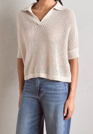 Lys beige strikket pullover med et takkede mesh-design, korte ærmer, v-hals og ribbet kant. Båret med blå højtaljede jeans.