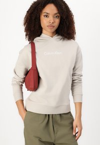 Béžová mikina s kapucňou na šnúrku a bielym logom "Calvin Klein", skombinovaná s olivovými voľnými nohavicami. Bordová kabelka cez plece dokončuje vzhľad.