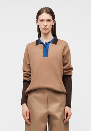 Kobieta ubrana w beżowy sweter z niebiesko-brązowym kołnierzem na guziki, na długim rękawie brązowej koszuli oraz pasujące beżowe spodnie, stojąca na białym tle.