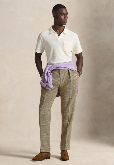 Άνδρας που στέκεται με τα χέρια στις τσέπες, φοράει πλεκτό πόλο κρεμ, καρό παντελόνι, καφέ μοκασίνια και λιλά πουλόβερ δεμένο γύρω από τη μέση.