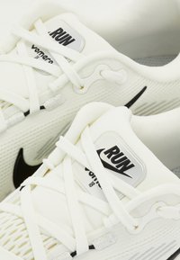 Hvide atletiske sko med en tekstureret mesh-overdel, sorte detaljer, flade snørebånd og en sølv logoetiket med teksten "RUN" og "Vomero."