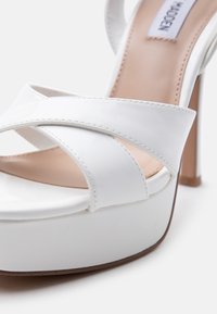 Λευκό δερμάτινο πέδιλο με ψηλό τακούνι και λουριά σε σχήμα χιαστί, με σόλα πλατφόρμα, εσωτερική επένδυση σε μπεζ χρώμα και εμφανείς ραφές.