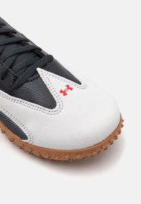 Scarpa sportiva bianca e nera con tomaia in pelle strutturata, suola in gomma con tacchetti e un logo rosso sul lato.