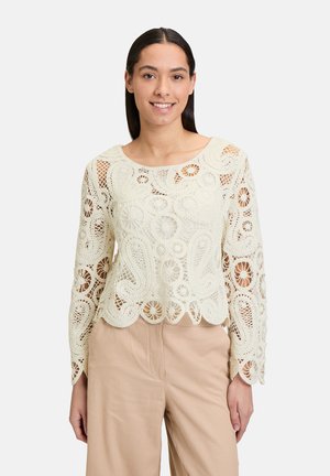 Top en dentelle couleur crème avec motifs floraux et paisley, manches longues et ourlet en feston, associé à un pantalon beige.