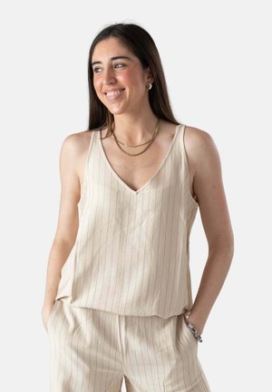 Joven mujer sonriendo, vestida con camiseta sin mangas de rayas beige y pantalones a juego, con collar y pulseras doradas, manos en los bolsillos, fondo blanco.