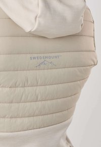 Dos de veste matelassée beige avec coutures horizontales et logo Swedemount représentant des montagnes et un oiseau en bleu-gris.