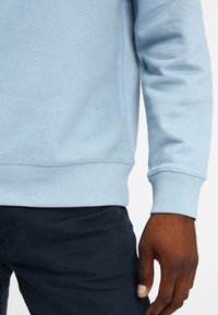 Höger arm och hand på en person som bär en ljusblå långärmad skjorta och mörka byxor mot en enfärgad bakgrund.