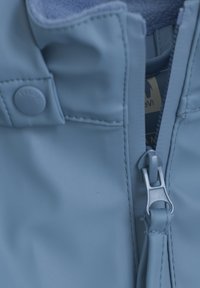 Ljust blå vattentät jacka med detalj av dragkedja och tryckknapp, med en mjuk insida synlig vid kragen.