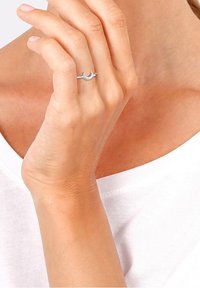 Main portant une bague en forme de croissant de lune argentée, touchant doucement le cou, avec une chemise blanche visible.