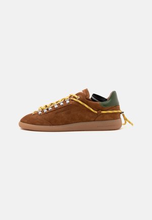 Baskets en daim marron avec lacets jaunes, languette de talon verte et semelle gomme, vue latérale sur fond blanc uni.