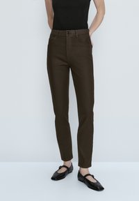 Vrouw in een donkerbruine slim-fit broek, een zwart mouwloos topje en zwarte Mary Jane flats, staand tegen een effen lichte achtergrond.