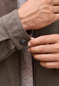 Brown leather jacket with a visible cuff, featuring a button with "SIGNATURE CO." text. The texture appears smooth, and the stitching is prominent.