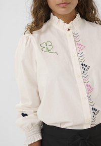 Blusa bianca con colletto a volant, maniche a sbuffo e vivaci motivi ricamati che includono un quadrifoglio verde e forme geometriche.
