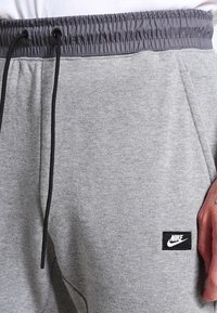 Šedé tepláky vyrobené z měkké tkaniny, s elastickým pasem, černým stahovacím páskem a malou černou nášivkou loga Nike na levém stehně.