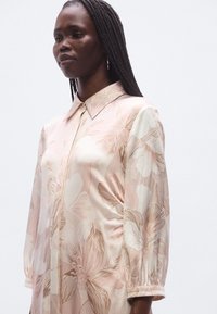 Φλοράλ φόρεμα από μεταξένιο ύφασμα με ανοιχτό ροζ φόντο και λευκά και χρυσά λουλουδένια σχέδια, γιακά με κουμπιά και μανίκια με μανσέτες.