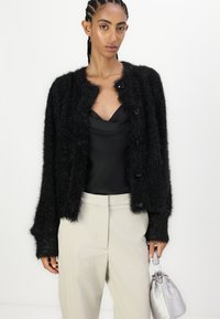 Fekete, bolyhos kardigán kerek nyakkivágással, gombos záródással és hosszú ujjakkal, selyem fekete toppal és bézs nadrággal párosítva. Ezüst táska.