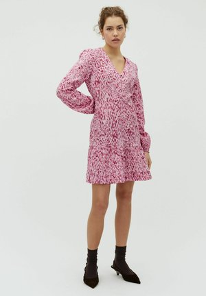 Mujer joven con cabello rizado que lleva un vestido de manga larga rosa con estampado, calcetas negras y zapatos negros de tacón bajo sobre un fondo blanco.