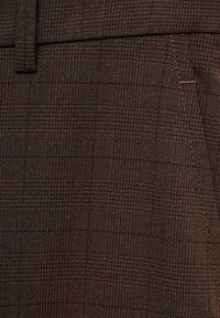 Hnědá tkanina se vzorem a texturovanou vazbou má mřížkový design, který zobrazuje jemné odstíny tmavší a světlejší hnědé barvy.