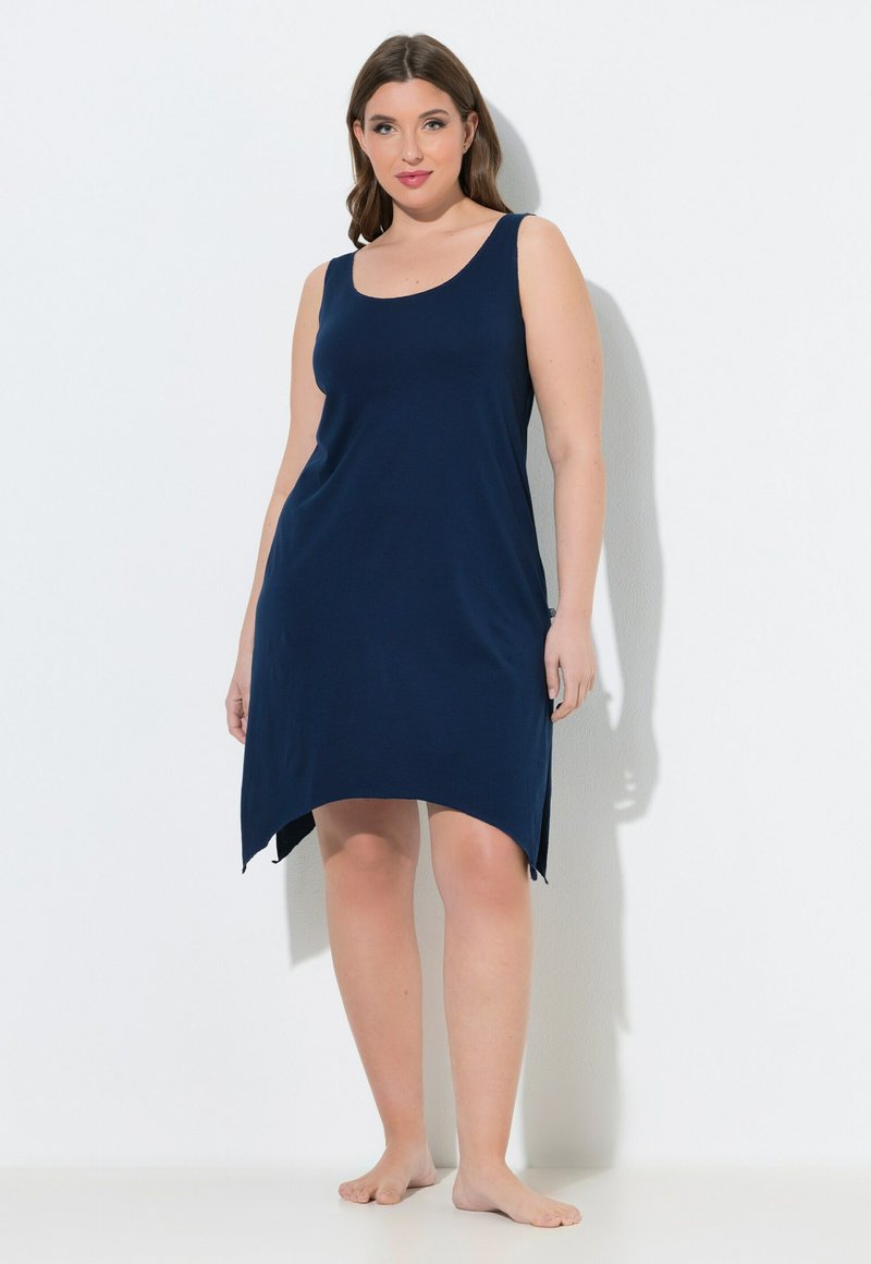 Mujer está descalza contra una pared blanca vistiendo un vestido azul marino sin mangas con un dobladillo desigual.