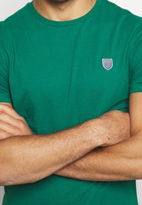 Grön bomullströja med rund halsringning och korta ärmar. Har en liten, broderad sköldlogotyp i vitt och marinblått.