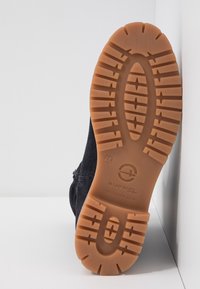 Semelle d'une botte noire avec une semelle en caoutchouc marron foncé et le logo de la marque, inclinée contre un fond blanc uni en angle.