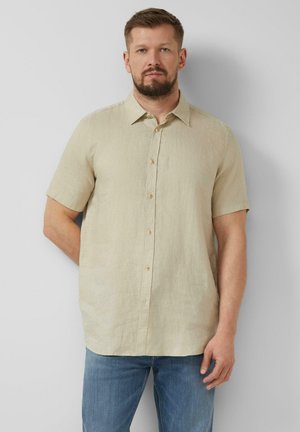 Mann mit Bart, der ein beiges kurzärmeliges Hemd und blaue Jeans trägt, steht vor einer einfarbigen hellgrauen Wand.