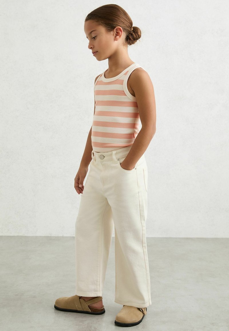 Jeune fille aux cheveux bruns attachés en chignon, portant un haut sans manches rayé rose et blanc, un pantalon blanc à jambes larges et des chaussures beiges sans lacets, debout de profil.