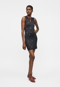 Kobieta w czarnej dopasowanej sukience bez rękawów z czerwonymi kwiatowymi wzorami i czerwonych balerinach na sznurowadła, stojąca z jedną ręką na biodrze.