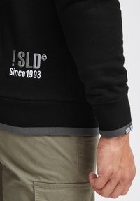 Crna dukserica s sivo obrubom, s izvezenim logotipom "! SLD Since 1993" sa strane. Tekstura tkanine i uski manžeti.