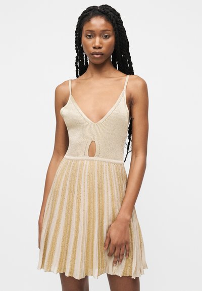 Vrouw met lang gevlochten haar die een beige gebreide jurk draagt met dunne bandjes, een diepe V-hals, een uitsnede aan de voorkant en een geplooide rok.