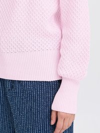 Hellrosa gestrickter Pullover mit einem strukturierten Muster, gerippten Bündchen und Saum. Getragen über blauen gestreiften Hosen mit vertikalen Linien.