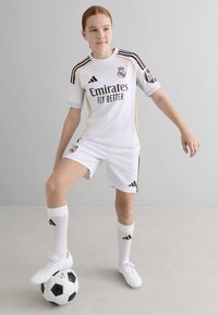 Balts futbola formas tērps ar melniem un zeltainiem akcentiem, ar Adidas zīmolu. Spēlētājs stāv uz melnbaltā futbola bumba. Baltas zeķes un apavi.