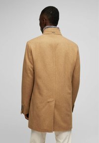 Abrigo de lana color camello con un corte estructurado, cuello alto y textura sutil, que presenta una sola abertura y puños abotonados.
