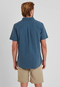 Short-sleeve blue button-up shirt with a back yoke detail, paired with khaki shorts. Cotton blend material, smooth texture.
