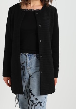 Femme portant un cardigan noir à manches longues boutonné sur un haut noir orné de dentelle, et un jean bleu clair avec un texte noir brodé.
