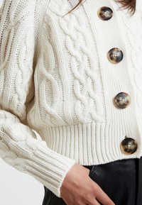 Cremefarbener Kabelstrickpullover mit gerippten Bündchen und Saum. Verfügt über große, runde, marmorierte Knöpfe entlang der Vorderseite. Weicher, strukturierter Stoff.