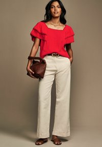 Blusa rossa con texture e maniche a volant, pantaloni beige a gamba larga e borsa intrecciata marrone. Presenta una fibbia rotonda in vita e sandali marroni.