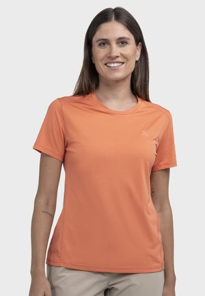 Lächelnde Frau mit glatten braunen Haaren, die ein oranges kurzärmeliges Shirt und beige Hosen vor einem schlichten hellen Hintergrund trägt.