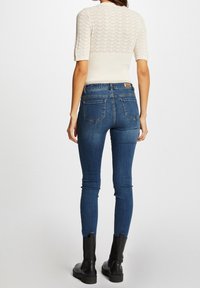 Häkeloberteil in Creme mit kurzen Ärmeln, kombiniert mit dunkelblauen Skinny Jeans. Schwarze Stiefeletten vervollständigen das Outfit. Die Jeans haben auf der Rückseite Taschen.