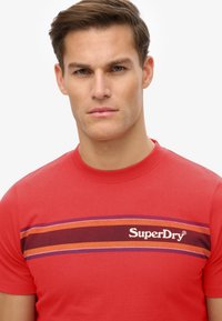 Rode katoenen T-shirt met horizontale strepen in oranje en paars, met het witte "SuperDry" logo. Ronde hals en korte mouwen.